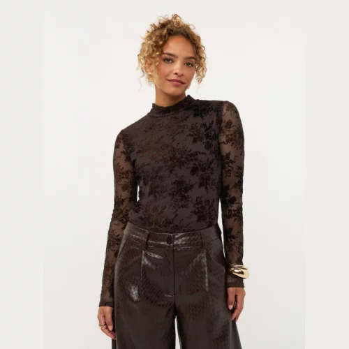 Model draagt Ydence top Brandy in donkerbruin met transparante uitstraling en lange mouwen, gecombineerd met een bruine pantalon.