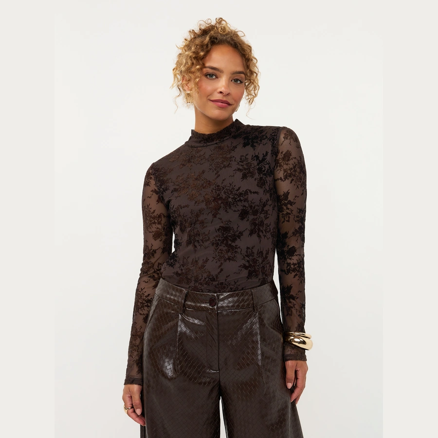 Model draagt Ydence top Brandy in donkerbruin met transparante uitstraling en lange mouwen, gecombineerd met een bruine pantalon.