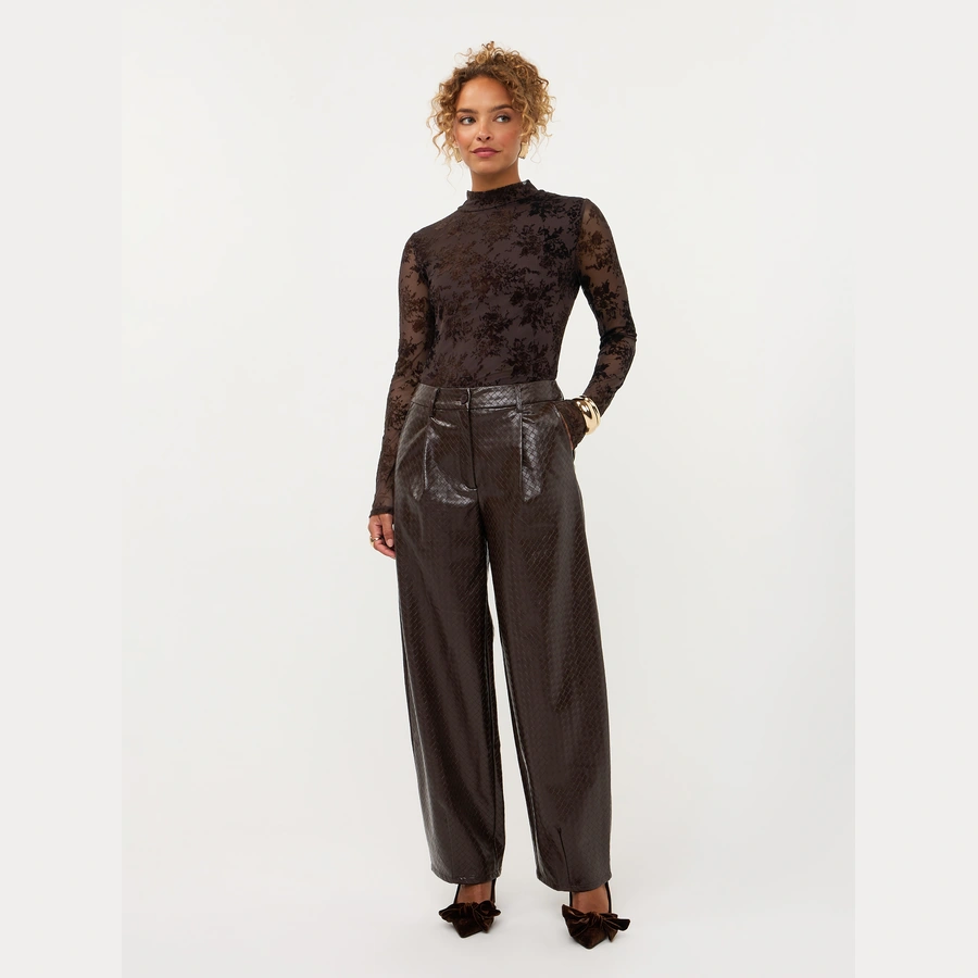Model draagt Ydence top Brandy in donkerbruin met transparante uitstraling en lange mouwen, gecombineerd met een bruine pantalon.